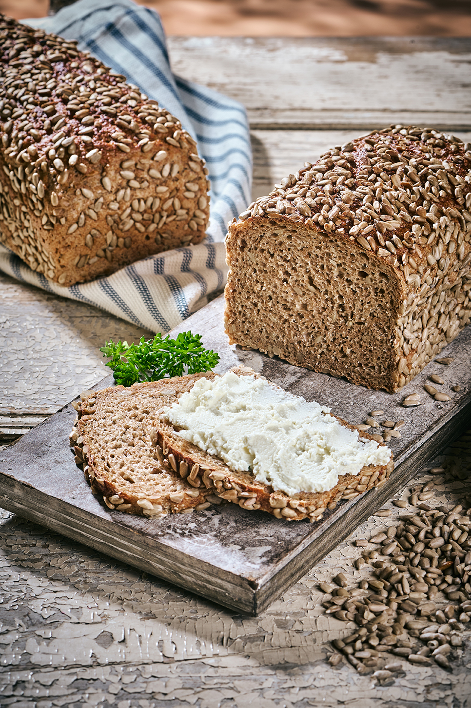 Dinkelvollkornbrot mit Sonnenblumenkernen und Frischkäse auf Holzbrett, daneben Körner.