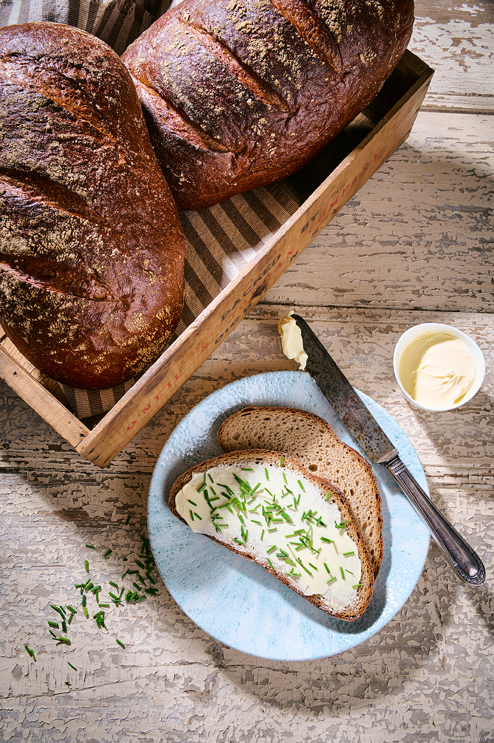 Roggenmischbrot in Scheiben mit Butter und Schnittlauch auf Teller, daneben ein Laib Brot in Kiste.