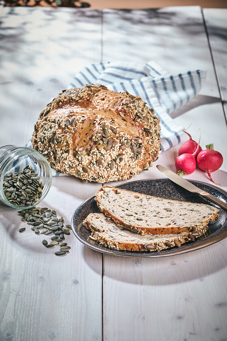 Kürbiskernquark-Brot mit Saaten, angeschnitten auf Holzbrett mit Radieschen und Kürbiskernen.