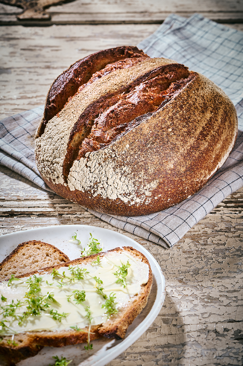 Dinkel-Steinofenbrot auf Küchentuch, Brotscheibe mit Butter und frischer Kresse auf Teller.
