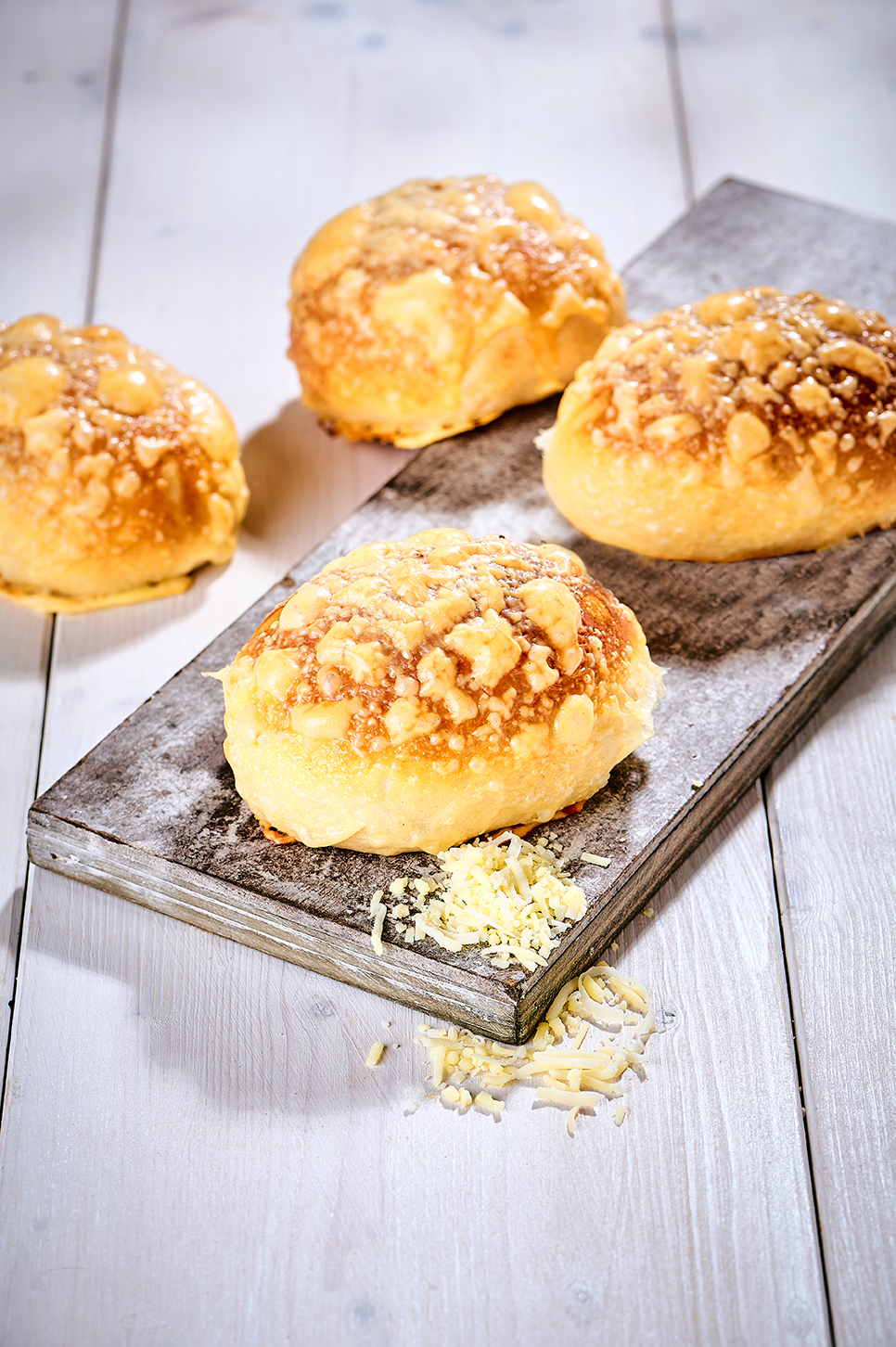 Vier frisch gebackene Käsebrötchen mit goldbrauner Käsekruste auf Holzbrett und Tisch.