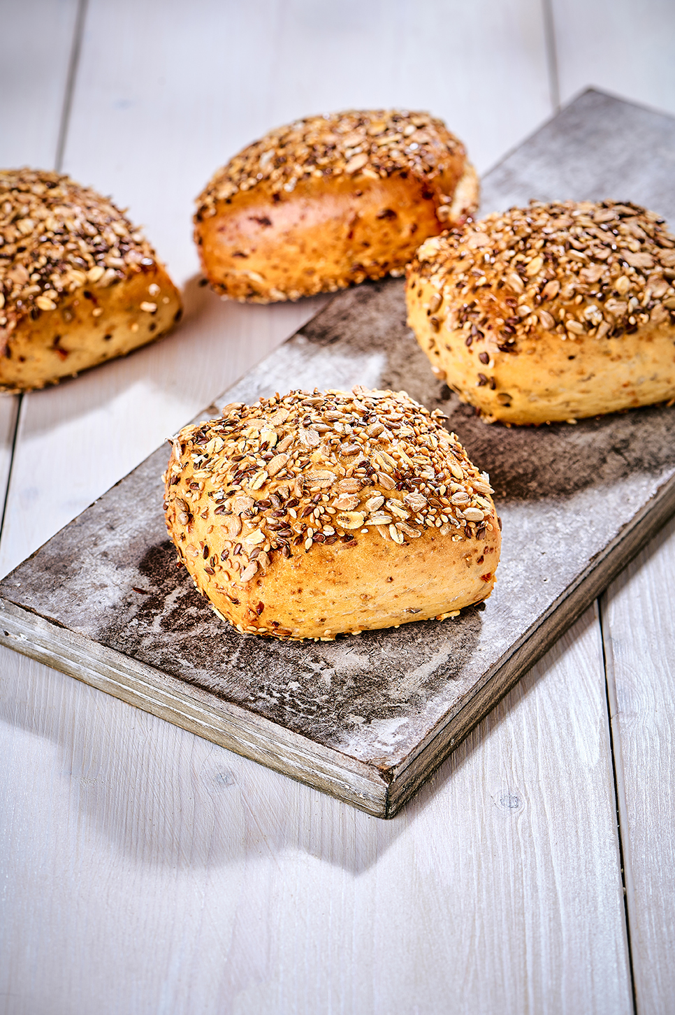 Vier Körnerbrötchen mit Saatenmischung auf bemehltem Holzbrett auf hellem Holztisch.