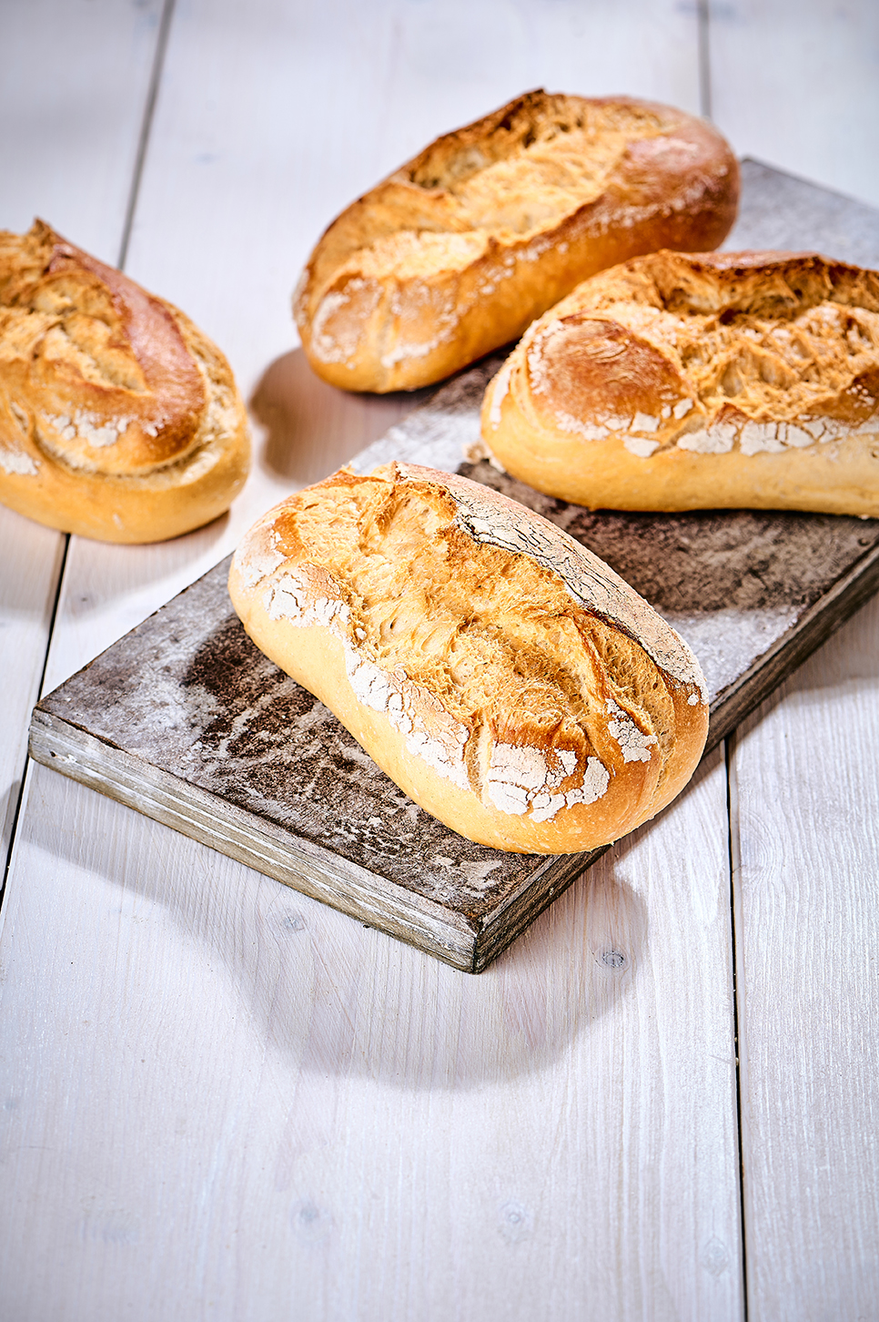 Vier rustikale Weizenbrötchen mit bemehlter Kruste auf Holzbrett auf hellem Holztisch.