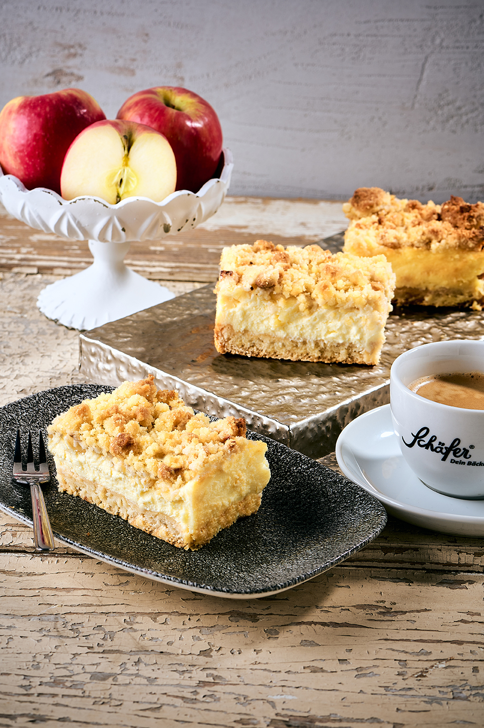 Stück Apfel-Streuselkuchen mit Kaffeetasse und frischen Äpfeln auf rustikalem Holztisch.