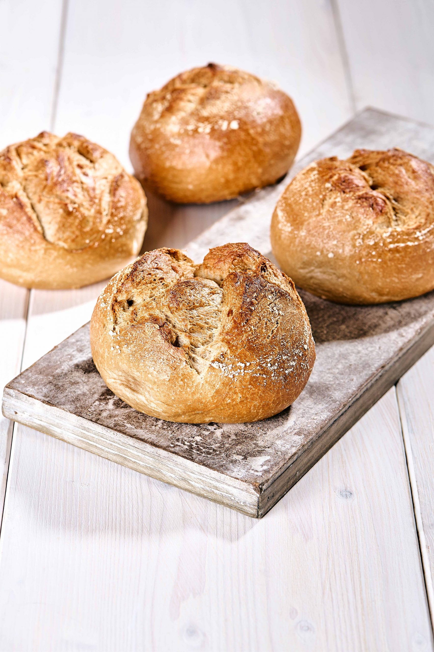 Vier rustikale Röggelchen mit bemehlter Kruste auf Holzbrett und hellem Holztisch.