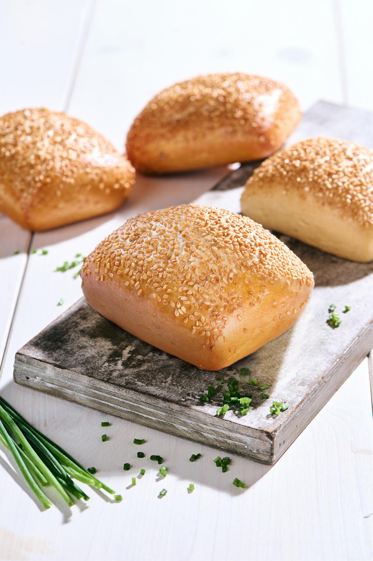 Vier goldbraune Sesambrötchen auf Holzbrett mit Schnittlauchstücken auf hellem Holztisch.